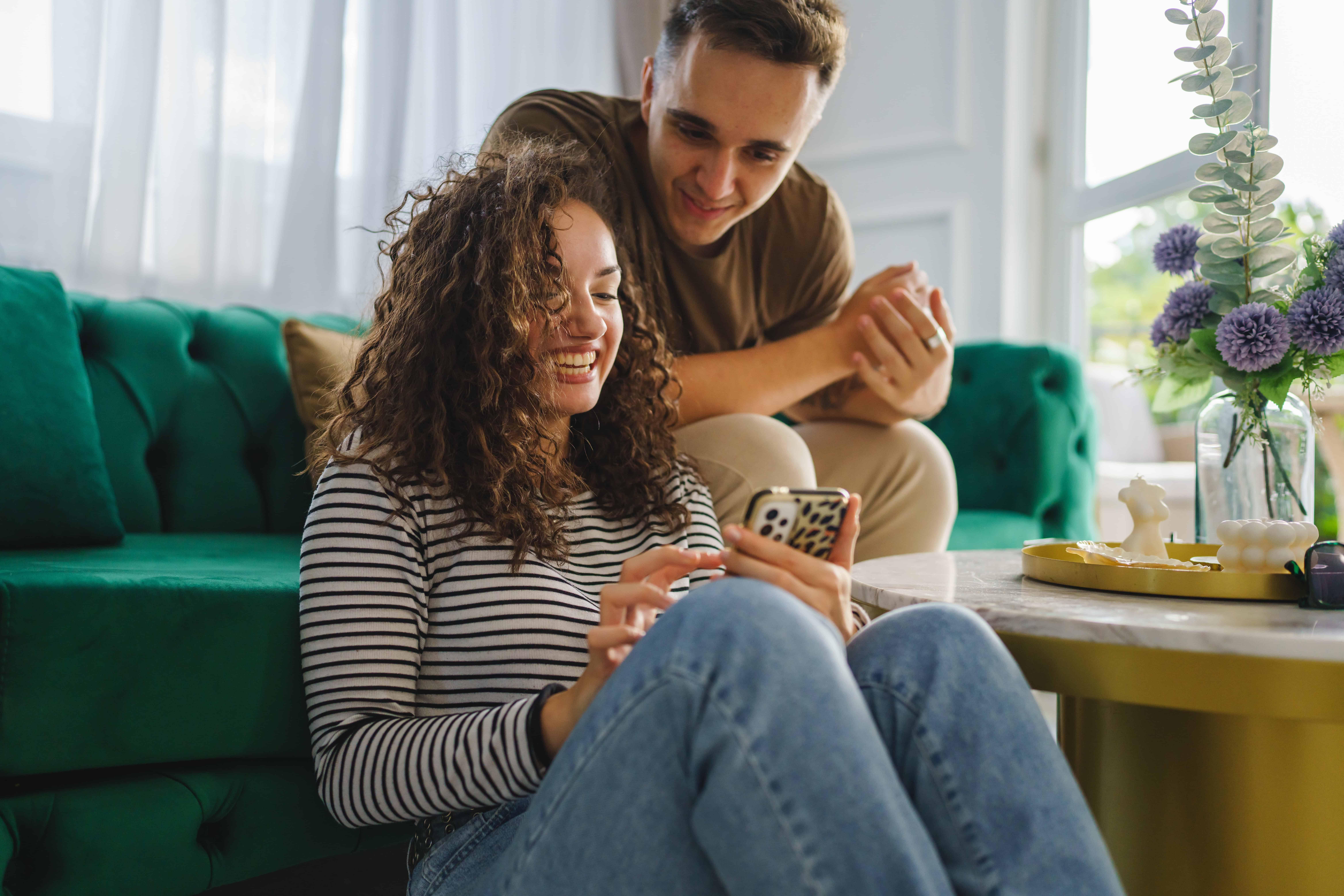 caucasian woman using mobile phone for online sms chat or internet browsing while her boyfriend or husband adult male sit beside at home in bright room real people couple leisure concept