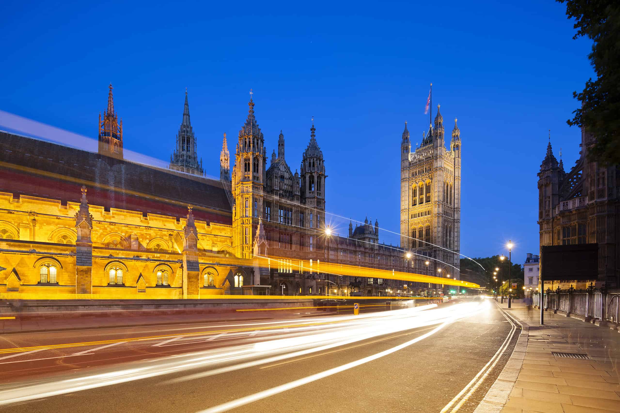 Long exposure night shot of the Houses of Parliament in London with blue sky and a street in the foreground.